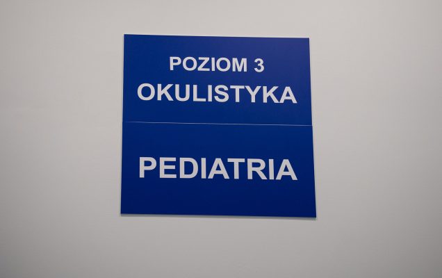 Dwie granatowe tablice informacyjne wewnątrz szpitala w Legionowie. Na pierwszej u g ory napis Poziom 3 Okulistyka, na drugiej poniżej napis Pediatria.
