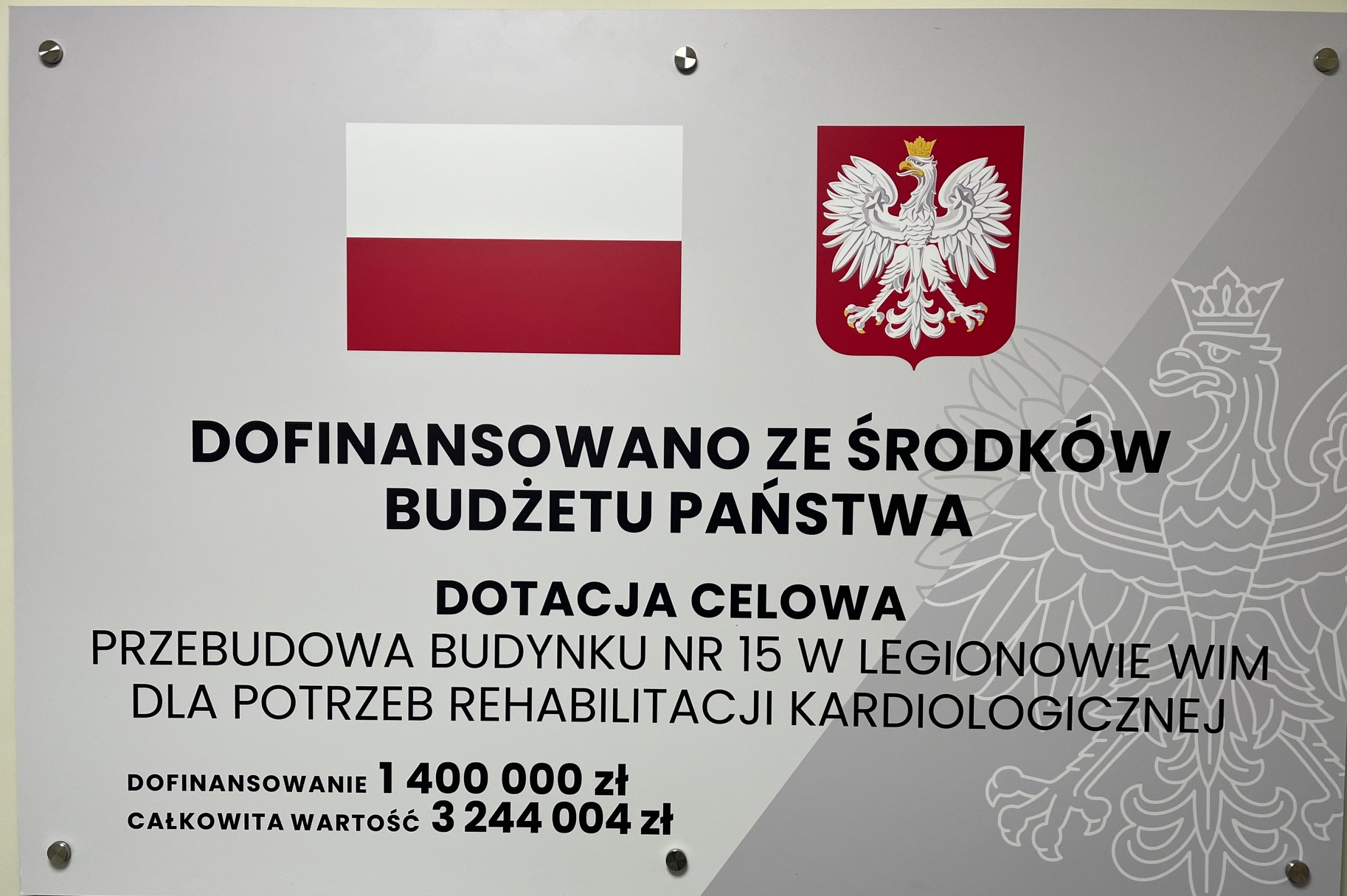 Tablica informacyjna dotacji celowej. Treść tablicy: Dofinansowano ze środków budżetu państwa, dotacja celowa. Przebudowa Budynku nr 15 w Legionowie WIM dla potrzeb rehabilitacji kardiologicznej. Dofinansowanie 1400 000 zł, całkowita wartość 3244 004 zł