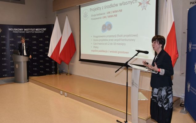 Uroczyste otwarcie Pracowni Anatomii w Wojskowym Instytucie Medycznym. Prelegenci przy mównicach po dwóch stronach sali, w tle slajd prezentacji.