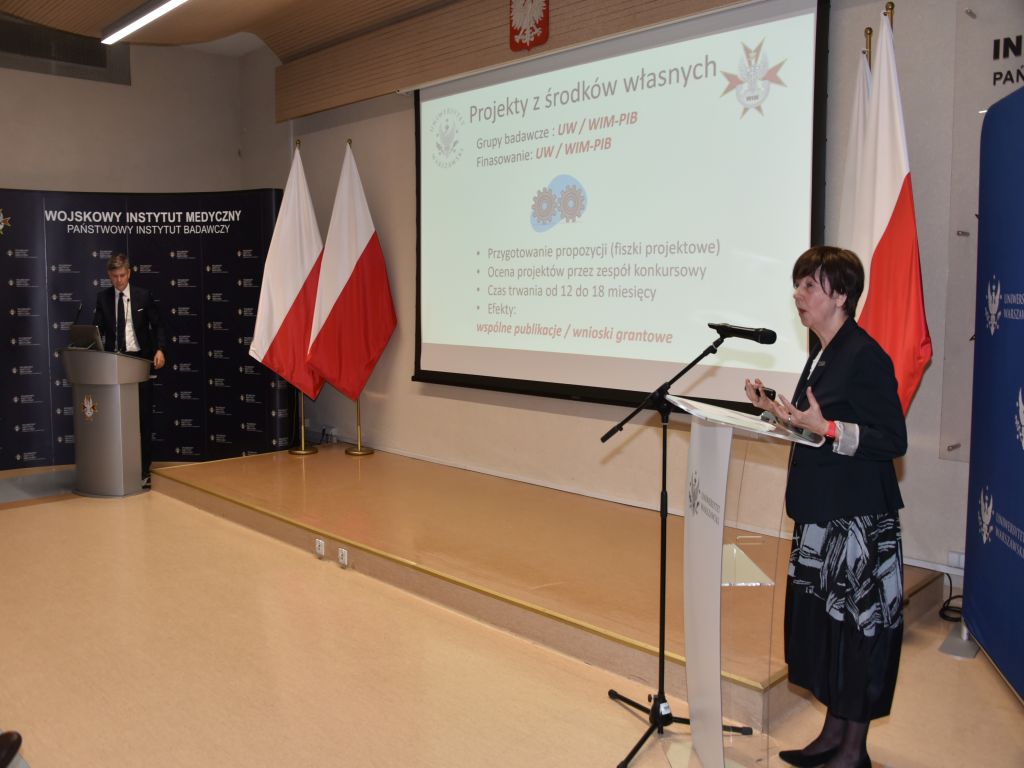 Uroczyste otwarcie Pracowni Anatomii w Wojskowym Instytucie Medycznym. Prelegenci przy mównicach po dwóch stronach sali, w tle slajd prezentacji.