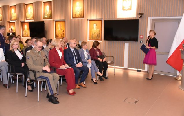 Małgorzata Złotkowska ubrana w różową sukienkę i czarny sweterek prowadzi uroczystość otwarcia nowej Poradni Chorób Piersi WIM-PIB. Na auli WIM siedzą zaproszeni goście. W pierwszym rzędzie dyrektor WIM Grzegorz Gielerak, Agata KORNHAUSER-DUDA