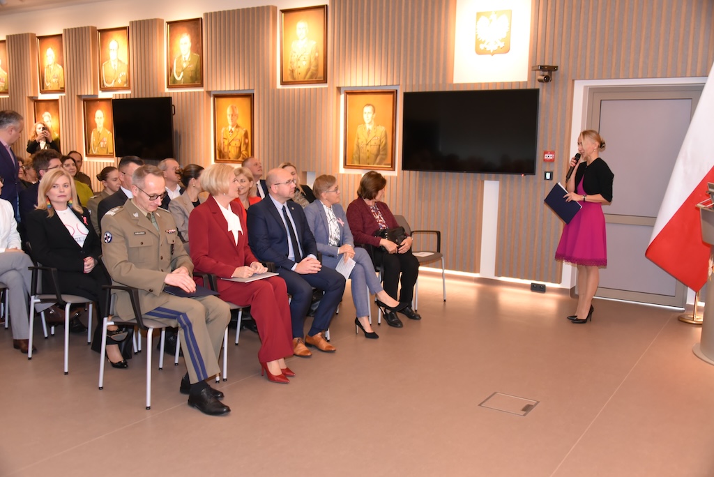 Małgorzata Złotkowska ubrana w różową sukienkę i czarny sweterek prowadzi uroczystość otwarcia nowej Poradni Chorób Piersi WIM-PIB. Na auli WIM siedzą zaproszeni goście. W pierwszym rzędzie dyrektor WIM Grzegorz Gielerak, Agata KORNHAUSER-DUDA