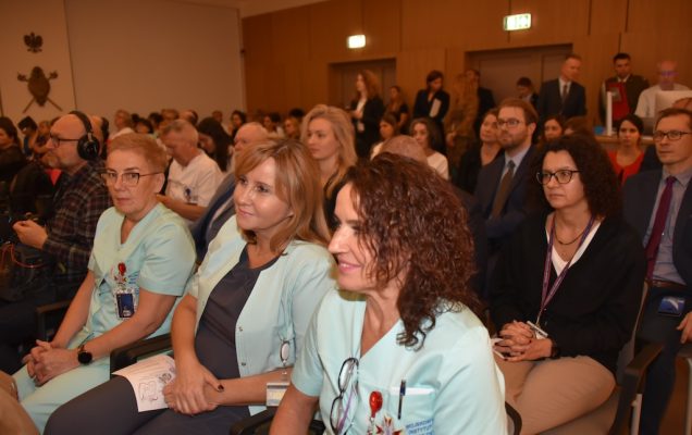Siedzący goście podczas uroczystości otwarcie nowej Poradni Chorób Piersi WIM-PIB. Na pierwszym planie pracownicy medyczni WIM w lekarskich uniformach, za nimi goście i pozostali pracownicy WIM.