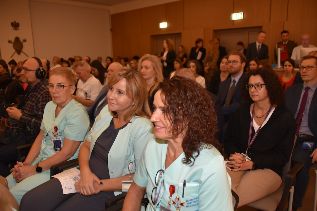 Siedzący goście podczas uroczystości otwarcie nowej Poradni Chorób Piersi WIM-PIB. Na pierwszym planie pracownicy medyczni WIM w lekarskich uniformach, za nimi goście i pozostali pracownicy WIM.