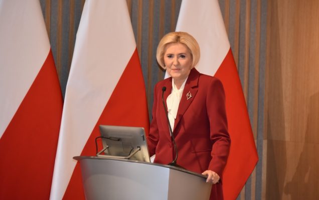 Agata KORNHAUSER-DUDA w czerwonym garniturze przy mównicy przemawia do zebranych gości, w tle dwie flagi Polski.