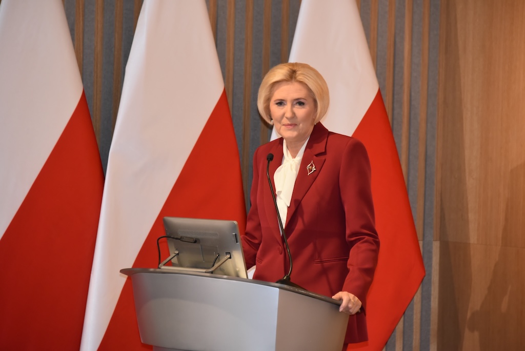 Agata KORNHAUSER-DUDA w czerwonym garniturze przy mównicy przemawia do zebranych gości, w tle dwie flagi Polski.