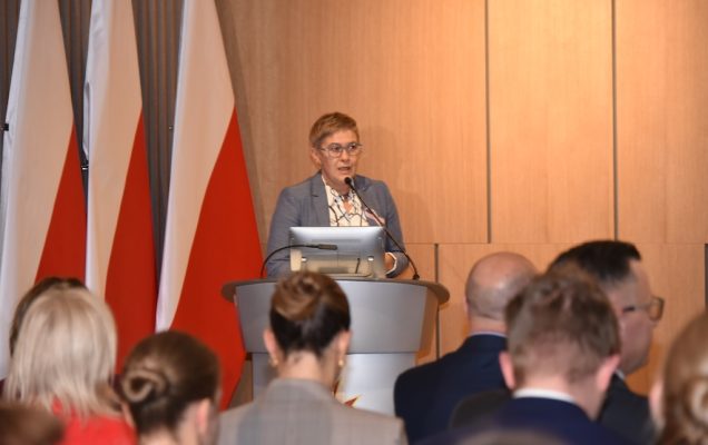 Podsekretarz stanu w MNiSW Maria Mrówczyńska ubrana w szary garnitur z przypiętą różową wstążeczką symbolizującą solidarność z chorymi na raka przemawia przy mównicy do zebranych gości, w tle dwie flagi Polski.