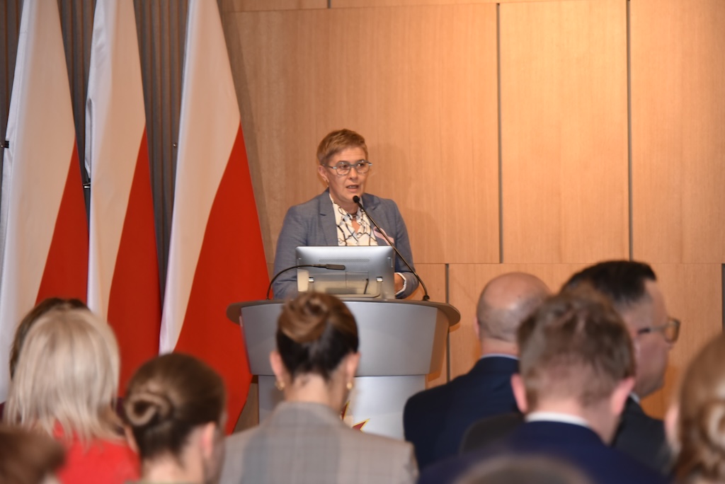 Podsekretarz stanu w MNiSW Maria Mrówczyńska ubrana w szary garnitur z przypiętą różową wstążeczką symbolizującą solidarność z chorymi na raka przemawia przy mównicy do zebranych gości, w tle dwie flagi Polski.