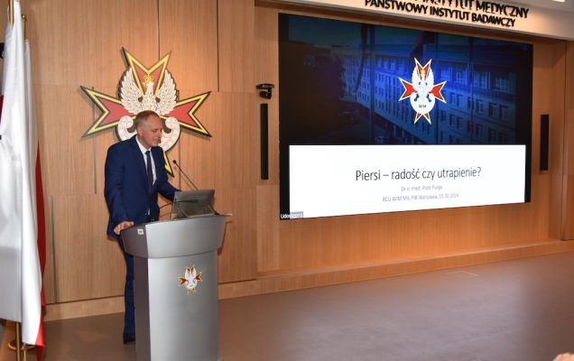 Doktor Piotr Furga stoi przy mównicy podczas wykładu. Na drugim planie za jego plecami wyświetlony fragment wykładu na którym wyświetlone logo WIM na granatowym tle oraz tytuł wykładu 