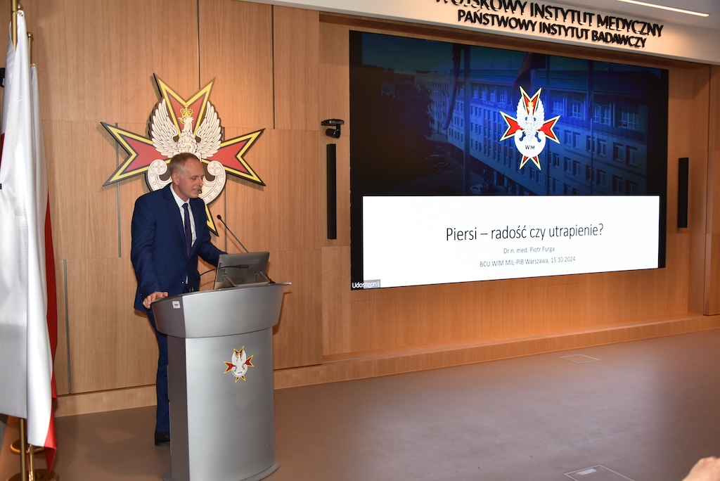 Doktor Piotr Furga stoi przy mównicy podczas wykładu. Na drugim planie za jego plecami wyświetlony fragment wykładu na którym wyświetlone logo WIM na granatowym tle oraz tytuł wykładu 