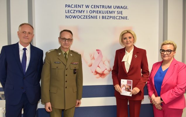 Korytarz Poradni Chorób Piersi. Po prawej Agata KORNHAUSER-DUDA prezentująca ulotkę Poradni Chorób Piersi obok Lidia Wojda, po lewej stronie dyrektor WIM Grzegorz Gielerak oraz Rzecznik Praw Pacjenta, Minister – Bartłomiej CHMIELOWIEC. Za nimi napis na ścianie 
