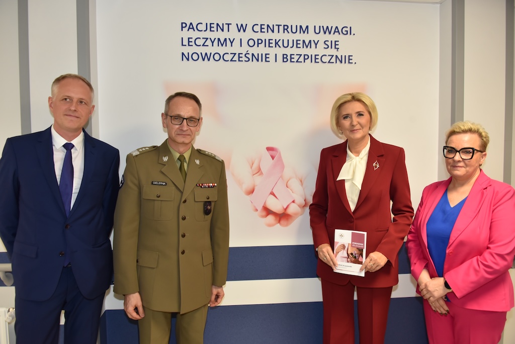 Korytarz Poradni Chorób Piersi. Po prawej Agata KORNHAUSER-DUDA prezentująca ulotkę Poradni Chorób Piersi obok Lidia Wojda, po lewej stronie dyrektor WIM Grzegorz Gielerak oraz Rzecznik Praw Pacjenta, Minister – Bartłomiej CHMIELOWIEC. Za nimi napis na ścianie 