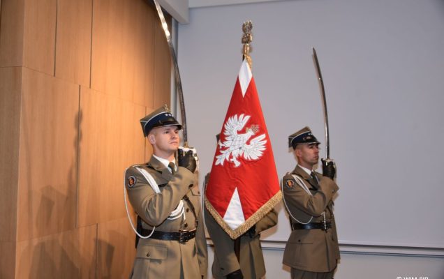 Relacja z Inauguracji Roku Akademickiego 2024/2025 w WIM-PIB. Trzej żołnierze ubrani w ceremonialne mundury, dwóch z nich stoi po bokach z uniesionymi szablami, środkowy trzyma uniesiony sztandar.