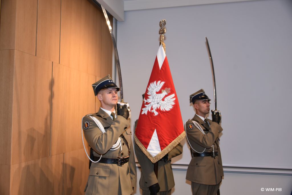 Relacja z Inauguracji Roku Akademickiego 2024/2025 w WIM-PIB. Trzej żołnierze ubrani w ceremonialne mundury, dwóch z nich stoi po bokach z uniesionymi szablami, środkowy trzyma uniesiony sztandar.