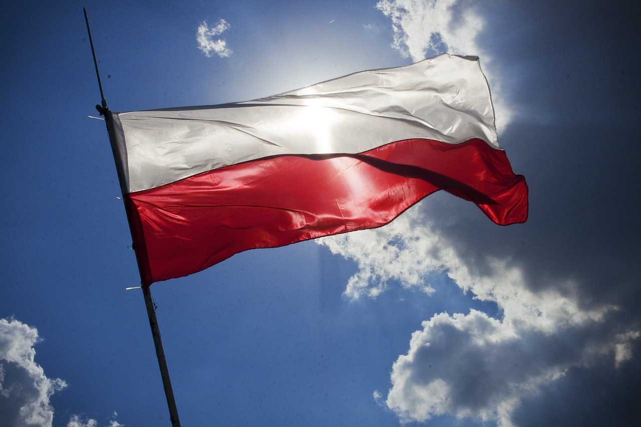 Flaga Polski na tle nieba, przez nią prześwituje słońce.