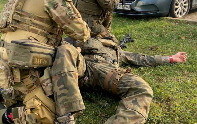 Żołnierze opatrujący rany podczas symulowanych ćwiczeń ratunkowych w wojskowym instytucie medycznym w warszawie. Jeden z żołnierzy opatruje leżącego na trawniku żołnierza. TRzeci żołnierz z karabinem w ręku klęczy odwrócony plecami do nich i zabezpiecza teren.