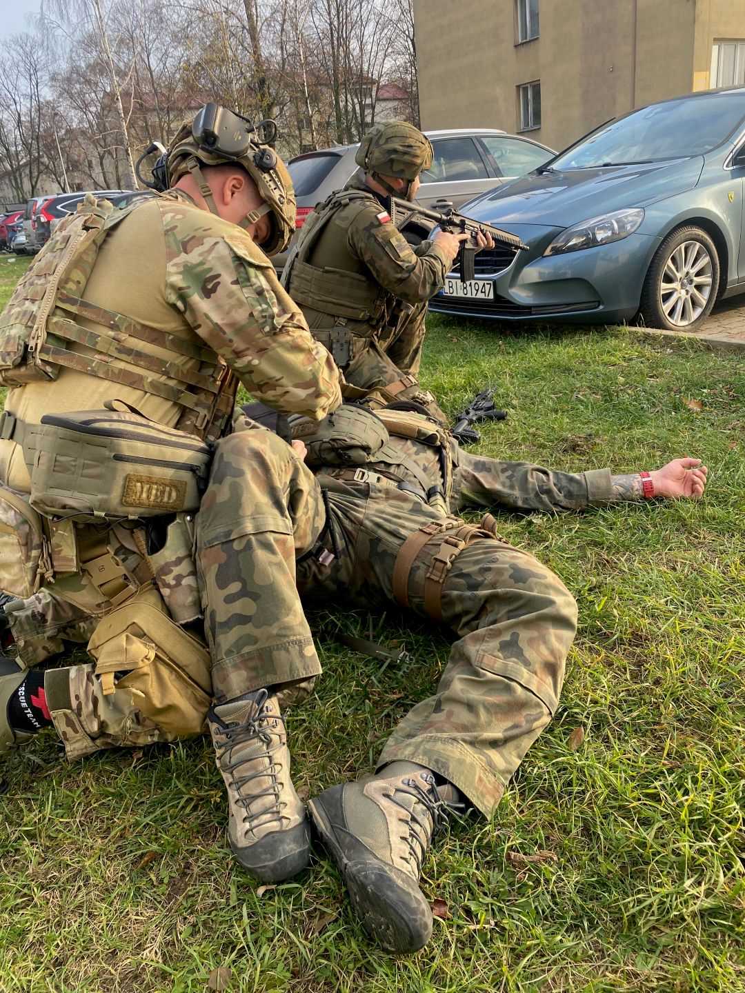 Żołnierze opatrujący rany podczas symulowanych ćwiczeń ratunkowych w wojskowym instytucie medycznym w warszawie. Jeden z żołnierzy opatruje leżącego na trawniku żołnierza. TRzeci żołnierz z karabinem w ręku klęczy odwrócony plecami do nich i zabezpiecza teren.