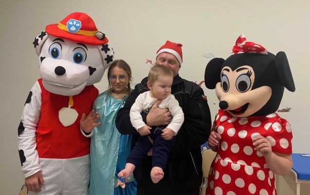 Mikołajki w Szpitalu w Legionowie. Korytarz szpitalny Goście z Ochotniczej Straży Pożarnej w Jadowie, przedstawiciele sołectwa Wólki Sulejowskiej, członków Koła Łowieckiego Cyranka oraz Stowarzyszenia Gospodyń Wiejskich w Myszadłach przebrani za postacie z kreskówki. Po środku członek ochotniczej straży pożarnej trzyma na rękach maleńkie dziecko.