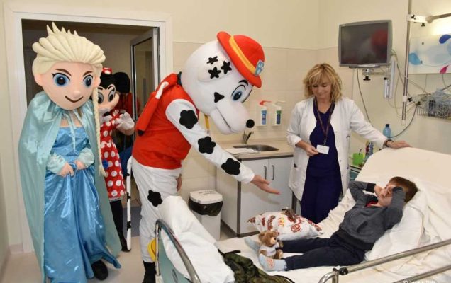 Zdjęcie kolorowe. Mikołajki w Wojskowym Instytucie Medycznym. Klinika Pediatryczna. Postaci z bajek Psi patrol i Kraina lodu witają się z chłopcem leżącym na łóżku w sali szpitalnej. Chłopiec zakrywa twarz rączkami, obok stoi pani pielęgniarka z kliniki pediatrycznej.