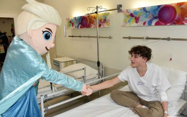 Zdjęcie kolorowe. Mikołajki w Wojskowym Instytucie Medycznym. Klinika Pediatryczna. Postać z kreskówki Kraina Lodu wita się z nastoletnim chłopcem siedzącym na łóżku szpitalnym.