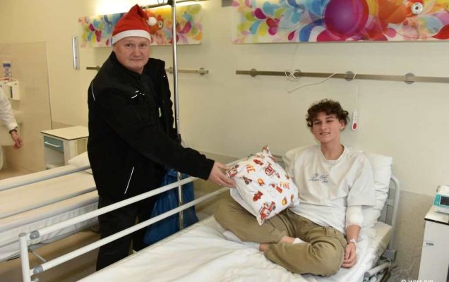 Zdjęcie kolorowe. Mikołajki w Wojskowym Instytucie Medycznym. Klinika Pediatryczna. Nastoletnim chłopiec siedzi na łóżku szpitalnym z poduszką prezentem obok funkcjonariusz ochotniczej straży pożarnej