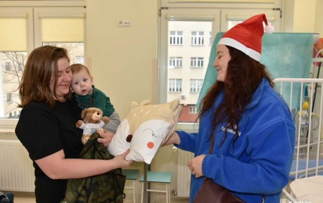 Zdjęcie kolorowe. Mikołajki w Wojskowym Instytucie Medycznym. Klinika Pediatryczna. Mama z niemowlakiem w sali szpitalnej odbiera prezent poduszkę prezent od dziewczyny ubranej w czapkę mikołaja.