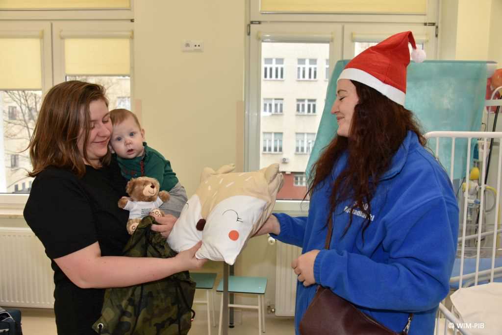Zdjęcie kolorowe. Mikołajki w Wojskowym Instytucie Medycznym. Klinika Pediatryczna. Mama z niemowlakiem w sali szpitalnej odbiera prezent poduszkę prezent od dziewczyny ubranej w czapkę mikołaja.