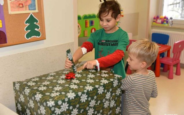 Zdjęcie kolorowe. Mikołajki w Wojskowym Instytucie Medycznym. Klinika Pediatryczna. Dwóch małych chłopców rozpakowuje duży prezent zapakowany w zielony świąteczny papier.