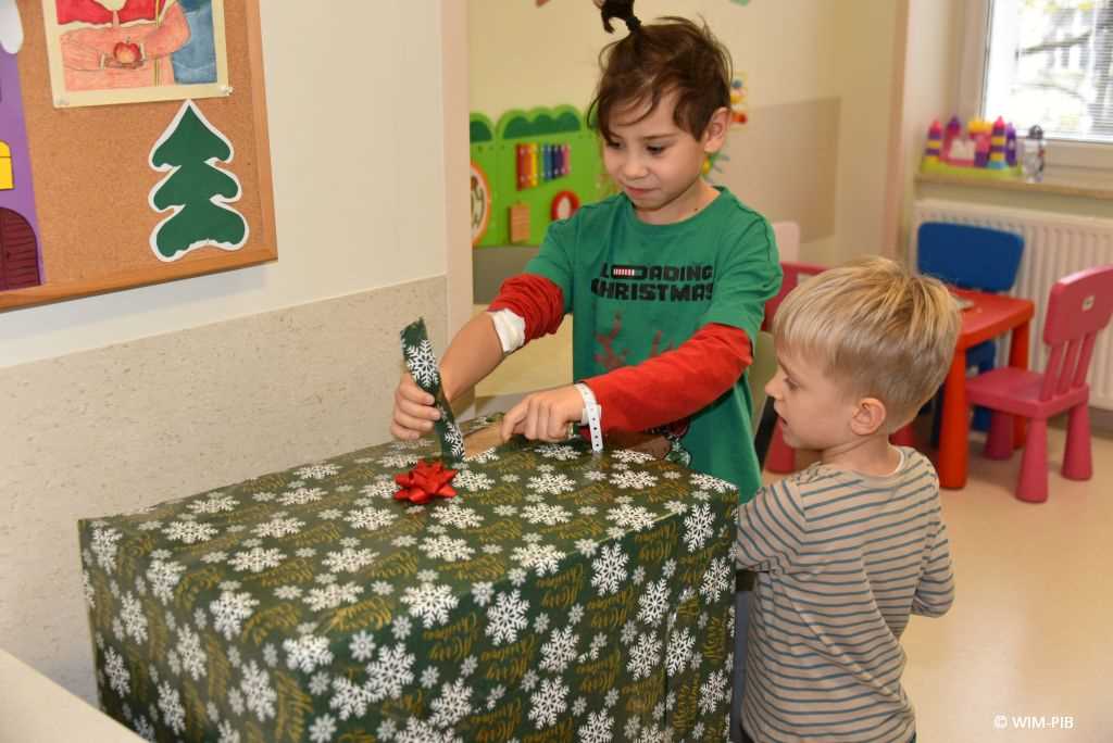 Zdjęcie kolorowe. Mikołajki w Wojskowym Instytucie Medycznym. Klinika Pediatryczna. Dwóch małych chłopców rozpakowuje duży prezent zapakowany w zielony świąteczny papier.