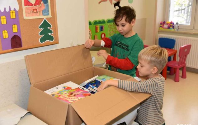 Zdjęcie kolorowe. Mikołajki w Wojskowym Instytucie Medycznym. Klinika Pediatryczna. Dwóch małych chłopców rozpakowuje duży prezent. W środku kartonu książeczki i zabawki.