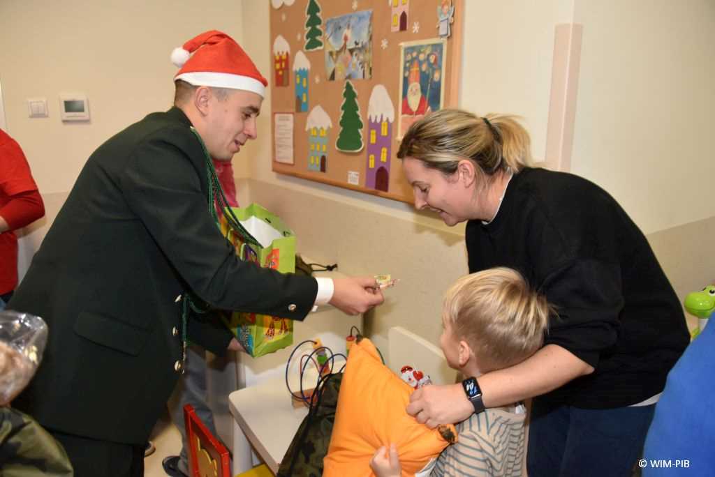 Zdjęcie kolorowe. Mikołajki w Wojskowym Instytucie Medycznym. Klinika Pediatryczna. Sala szpitalna. Chłopiec i obejmująca go mama odpierają prezent mikołajkowy od przedstawiciela ochotniczej straży pożarnej.