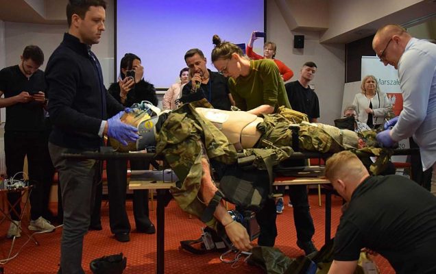 Pracownicy WIM biorą udział w ćwiczeniach medycznych. Na stole fantom w wojskowym ubraniu, wokoło niego grupa uczestników uczy się opatrywać rany.