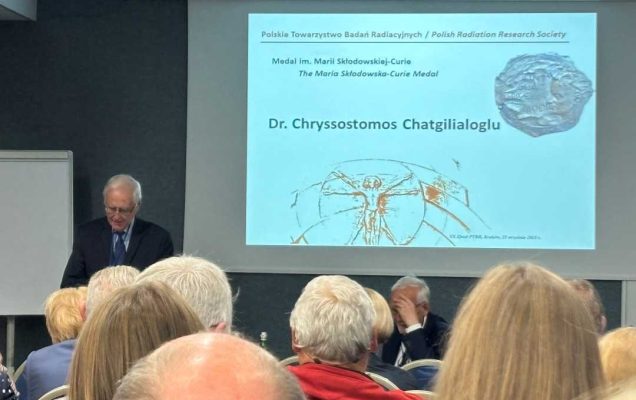 Sala wykładowa, ujęcie z tyłu sali uczestników siedzących na sali konferencyjnej podczas wykładu. Przed siedzącymi stojący prelegent przy prezentacji wyświetlanej z rzutnika.