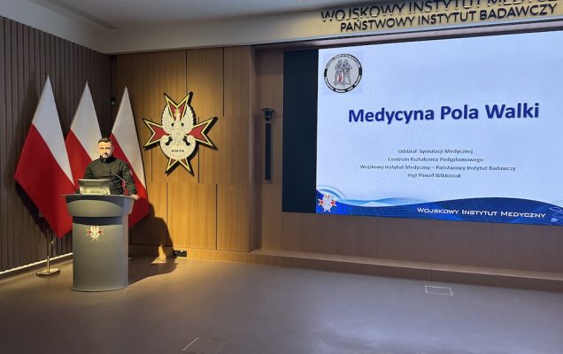 Prelegent przy mównicy podczas konferencji, za nim na wyświetlaczu prezentacja Medycyna Pola Walki.
