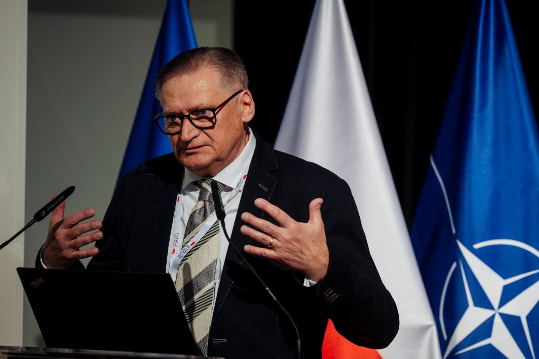 Prelegent przy mównicy. Za jego plecami flagi UE, Polski, NATO.