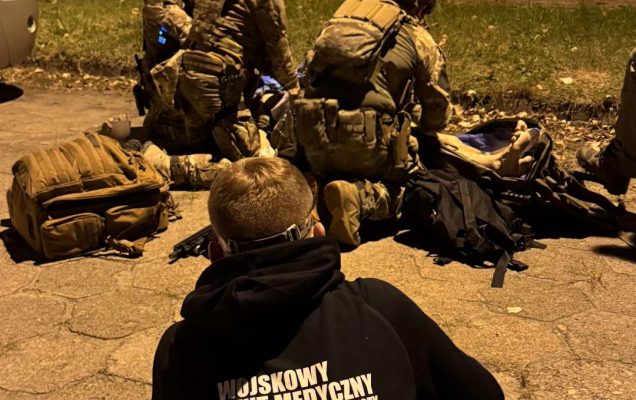 Ćwiczenia medyczne w plenerze. Trzech ratowników w strojach wojskowych klęczy nad poszkodowanym, na pierwszym planie za plecami ratowników kucający młody mężczyzna ubrany w bluzę z napisem na plecach Wojskowy Instytut Medyczny.