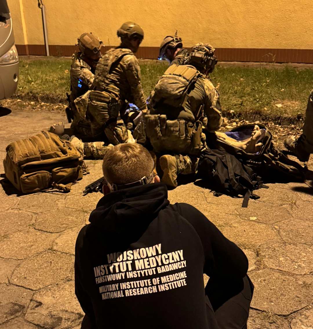 Ćwiczenia medyczne w plenerze. Trzech ratowników w strojach wojskowych klęczy nad poszkodowanym, na pierwszym planie za plecami ratowników kucający młody mężczyzna ubrany w bluzę z napisem na plecach Wojskowy Instytut Medyczny.