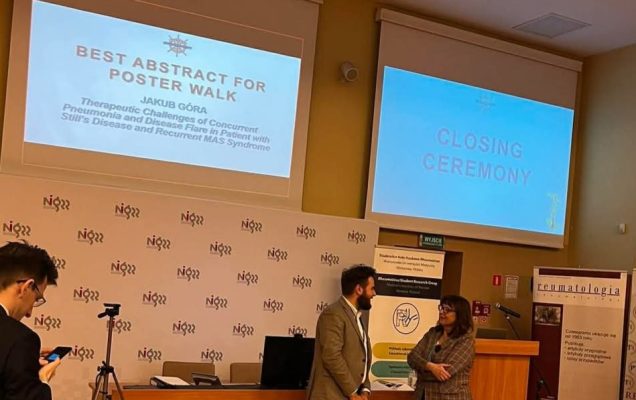 Sala wykładowa podczas ceremonii zamknięcia konferencji naukowej. Na dwóch ekranach widoczne są slajdy: po lewej napis „Best Abstract for Poster Walk – Jakub Góra” wraz z tytułem pracy „Therapeutic Challenges of Concurrent Pneumonia and Disease Flare in Patient with Still’s Disease and Recurrent MAS Syndrome”, a po prawej – „Closing Ceremony”. Na dole zdjęcia widać podium i biurko z komputerem, przed którymi stoją dwie osoby w rozmowie – mężczyzna w garniturze i kobieta w kraciastej marynarce. Po lewej stronie osoba z telefonem i statywem, a w tle banery z logo Warszawskiego Uniwersytetu Medycznego, Studenckiego Koła Naukowego Reumatologii oraz czasopisma „Reumatologia”.
