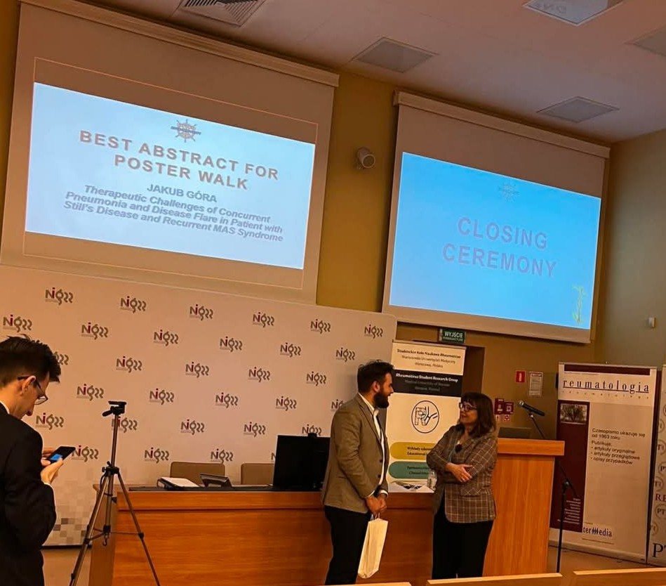 Sala wykładowa podczas ceremonii zamknięcia konferencji naukowej. Na dwóch ekranach widoczne są slajdy: po lewej napis „Best Abstract for Poster Walk – Jakub Góra” wraz z tytułem pracy „Therapeutic Challenges of Concurrent Pneumonia and Disease Flare in Patient with Still’s Disease and Recurrent MAS Syndrome”, a po prawej – „Closing Ceremony”. Na dole zdjęcia widać podium i biurko z komputerem, przed którymi stoją dwie osoby w rozmowie – mężczyzna w garniturze i kobieta w kraciastej marynarce. Po lewej stronie osoba z telefonem i statywem, a w tle banery z logo Warszawskiego Uniwersytetu Medycznego, Studenckiego Koła Naukowego Reumatologii oraz czasopisma „Reumatologia”.