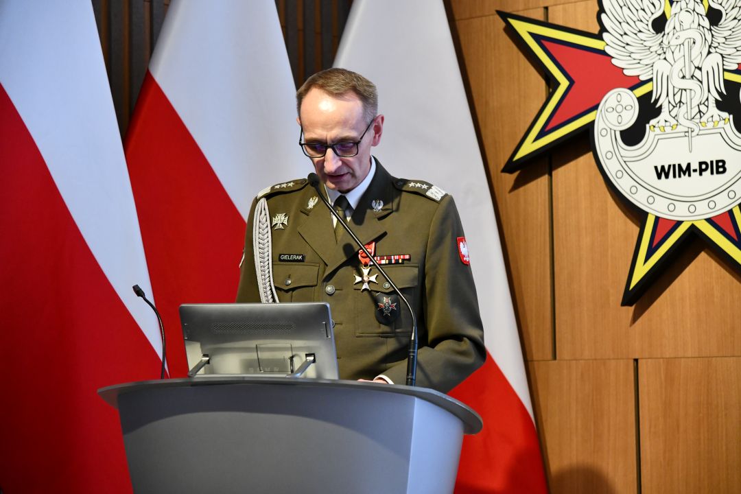 Dyrektor WIM przy mównicy, za nim trzy flagi polski i logo WIM.