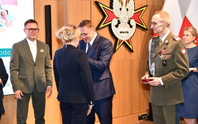 Mateusz Kotecki przypina kobiecie odznaczenie, obok stoi dyrektor WIM.