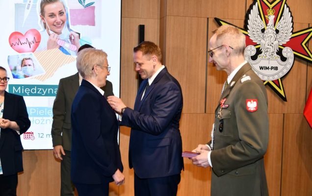 Mateusz Kotecki przypina kobiecie odznaczenie, obok stoi dyrektor WIM.