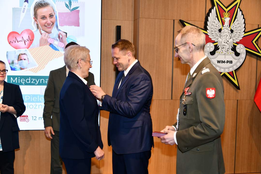 Mateusz Kotecki przypina kobiecie odznaczenie, obok stoi dyrektor WIM.