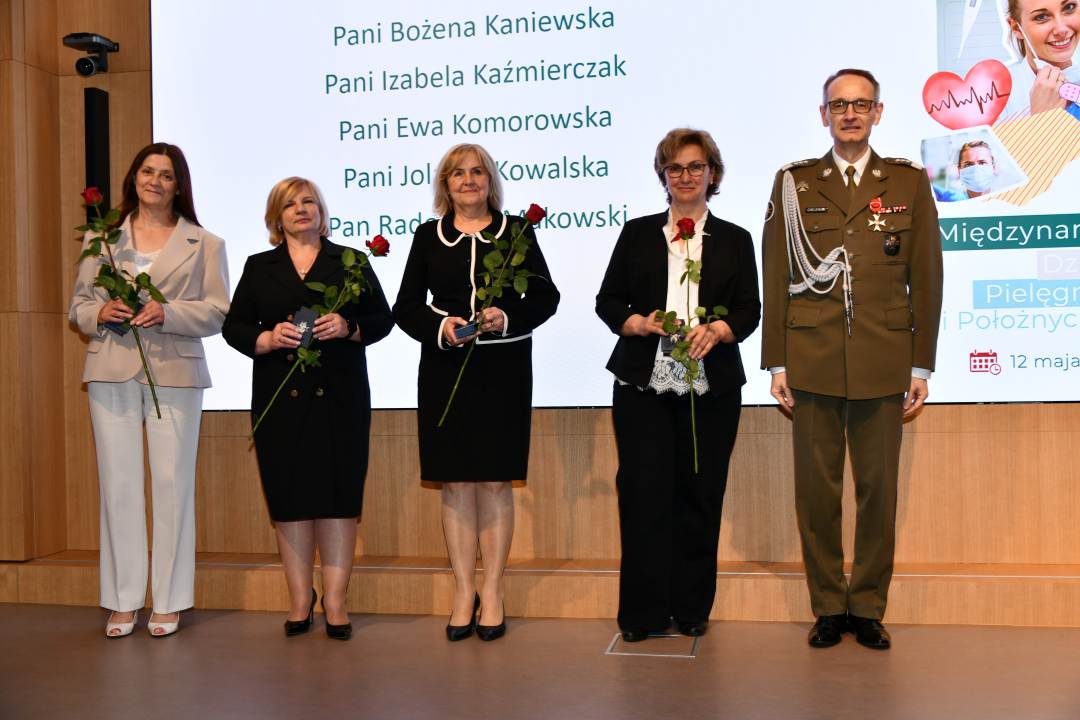 Dyrektor WIM pozuje do wspólnego zdjęcia z pielęgniarkami trzymającymi czerwone róże i odznaczenia.
