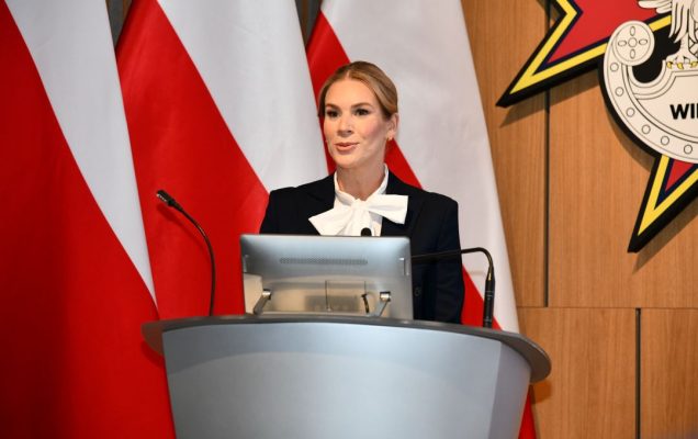 Pierwsza Dama RP przy mównicy, za jej plecami flaga Polski.