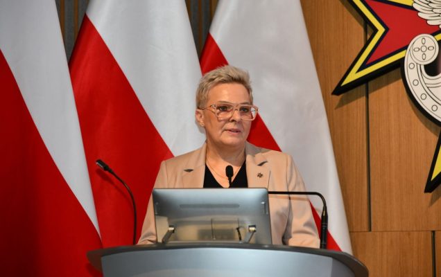 Lidja Wojda przy mównicy za jej plecami trzy flagi polski.