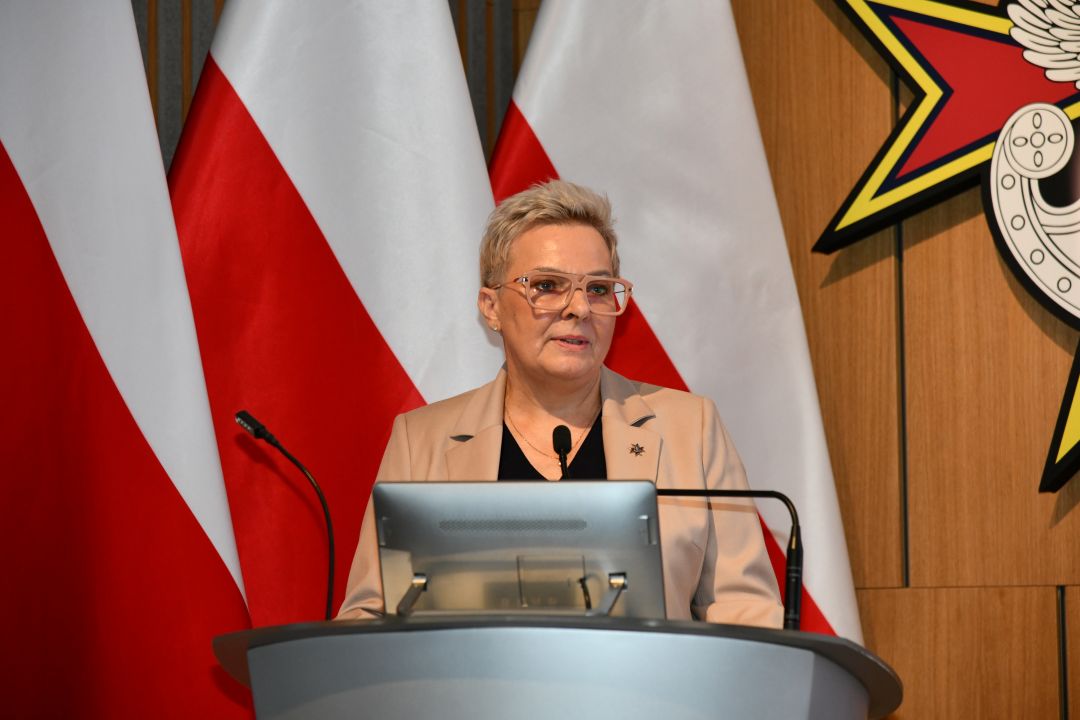 Lidja Wojda przy mównicy za jej plecami trzy flagi polski.
