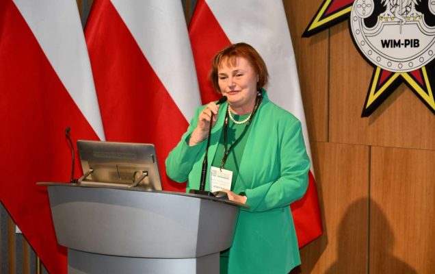 Zofia Sienkiewicz przy mównicy, za jej plecami trzy flagi Polski.