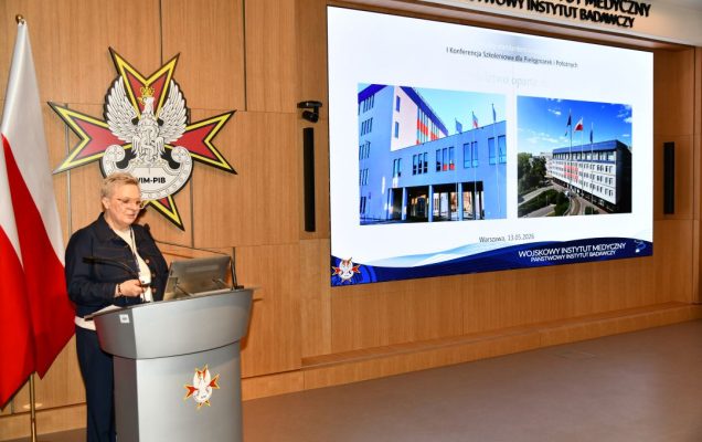 Lidia Wojda przy mównicy w sali Wojskowego Instytutu Medycznego; obok polska flaga i emblemat WIM‑PIB. Na ekranie widoczny slajd z tytułem ‘I Konferencja Szkoleniowa dla Pielęgniarek i Położnych’ oraz zdjęciami budynków instytutu.