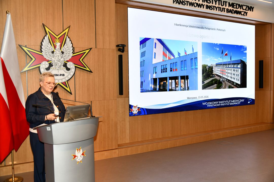 Lidia Wojda przy mównicy w sali Wojskowego Instytutu Medycznego; obok polska flaga i emblemat WIM‑PIB. Na ekranie widoczny slajd z tytułem ‘I Konferencja Szkoleniowa dla Pielęgniarek i Położnych’ oraz zdjęciami budynków instytutu.